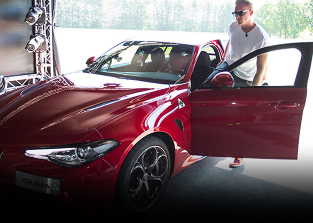Man in sunglasses beside red Alfa Romeo.